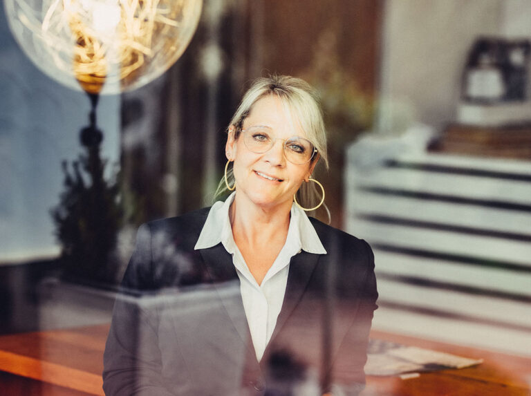 Frau mit blonden Haaren und Brille, sitzt in einem modernen Büro mit reflektierendem Fenster und einer großen Glühbirne im Vordergrund.