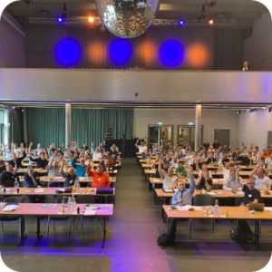 Großer Konferenzraum mit Teilnehmern an langen Tischen, beleuchtet von einer Discokugel an der Decke.