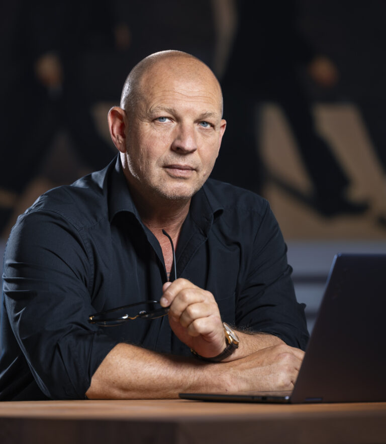 Geschäftsmann mit ernstem Blick, sitzt an einem Holztisch mit Laptop, hält eine Brille in der Hand.
