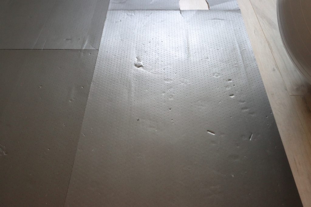 Unebene Estrichfläche mit verlegter Dämmunterlage, auf der punktuelle Erhebungen, Dellen und Druckstellen sichtbar sind. Die Unebenheiten zeigen die Folgen von Schmutz- und Mörtelresten unter der Unterlage, die zu lokalen Höhendifferenzen führen können.